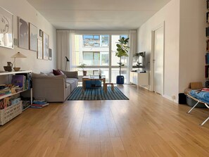 Ein Wohnzimmer mit einer Couch, einem Couchtisch und einem Fernseher. Es gibt Bücherregale und einen Blumentopf.
