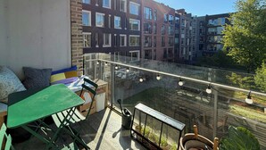 Un balcon doté d’une balustrade en verre, une table verte et des chaises, d’où l’on peut admirer une rangée d’immeubles à plusieurs étages.