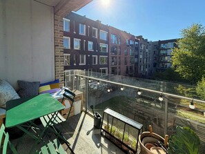 Ein Balkon mit Glastrennwand, einem grünen Tisch und Stühlen, mit Blick auf eine Reihe mehrstöckiger Gebäude.