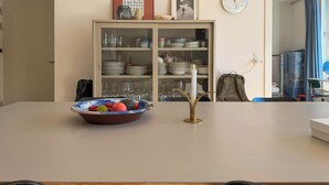 Un coin repas avec une table blanche, un saladier de fruits, une bougie allumée et un buffet contenant de la vaisselle.