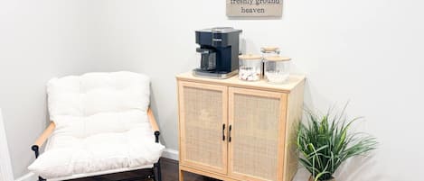 Ein Zimmer mit einem weißen, gepolsterten Sessel, einem Holzkabinett mit Kaffeemaschine und einem Blumentopf.