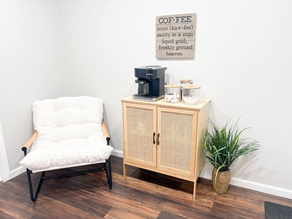 A room with a white cushioned chair, a wooden cabinet with a coffee maker, and a potted plant.