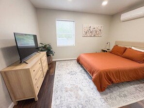 A bedroom with a bed, a TV on a wooden dresser, a window with blinds, and a wall-mounted air conditioner.