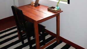 A room with a wooden table, a chair, a window with curtains, and a view of greenery outside.