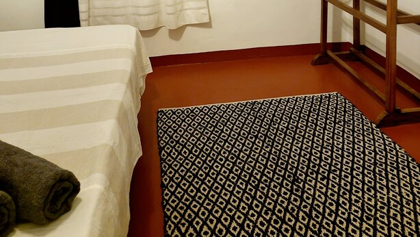 A bedroom with a bed, a wooden bunk bed, a window with sheer curtains, a patterned rug, and a wall-mounted mirror.