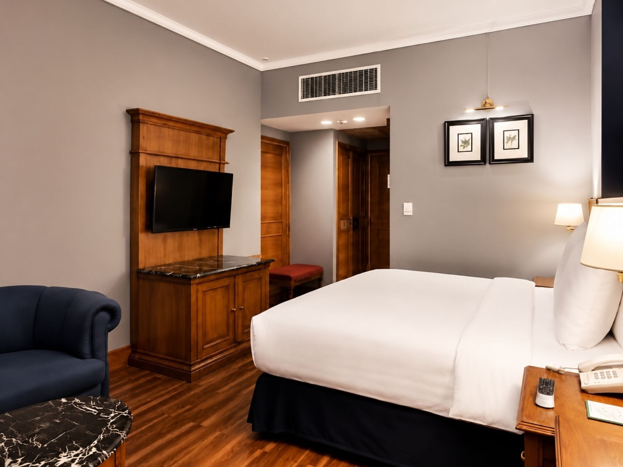 A hotel room with a bed, a TV mounted on a wooden cabinet, a desk with a lamp, and a chair.