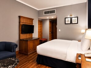 A hotel room with a bed, a TV mounted on a wooden cabinet, a desk with a lamp, and a chair.