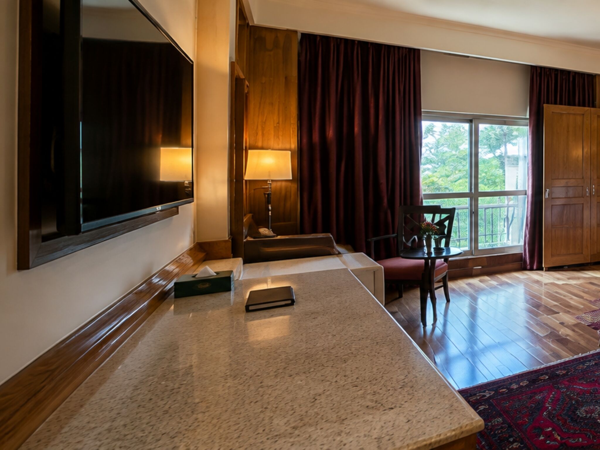 Quarto de hotel com uma TV de tela plana grande, um banco de madeira, uma pequena mesa com abajur e vista para o verde pela janela.