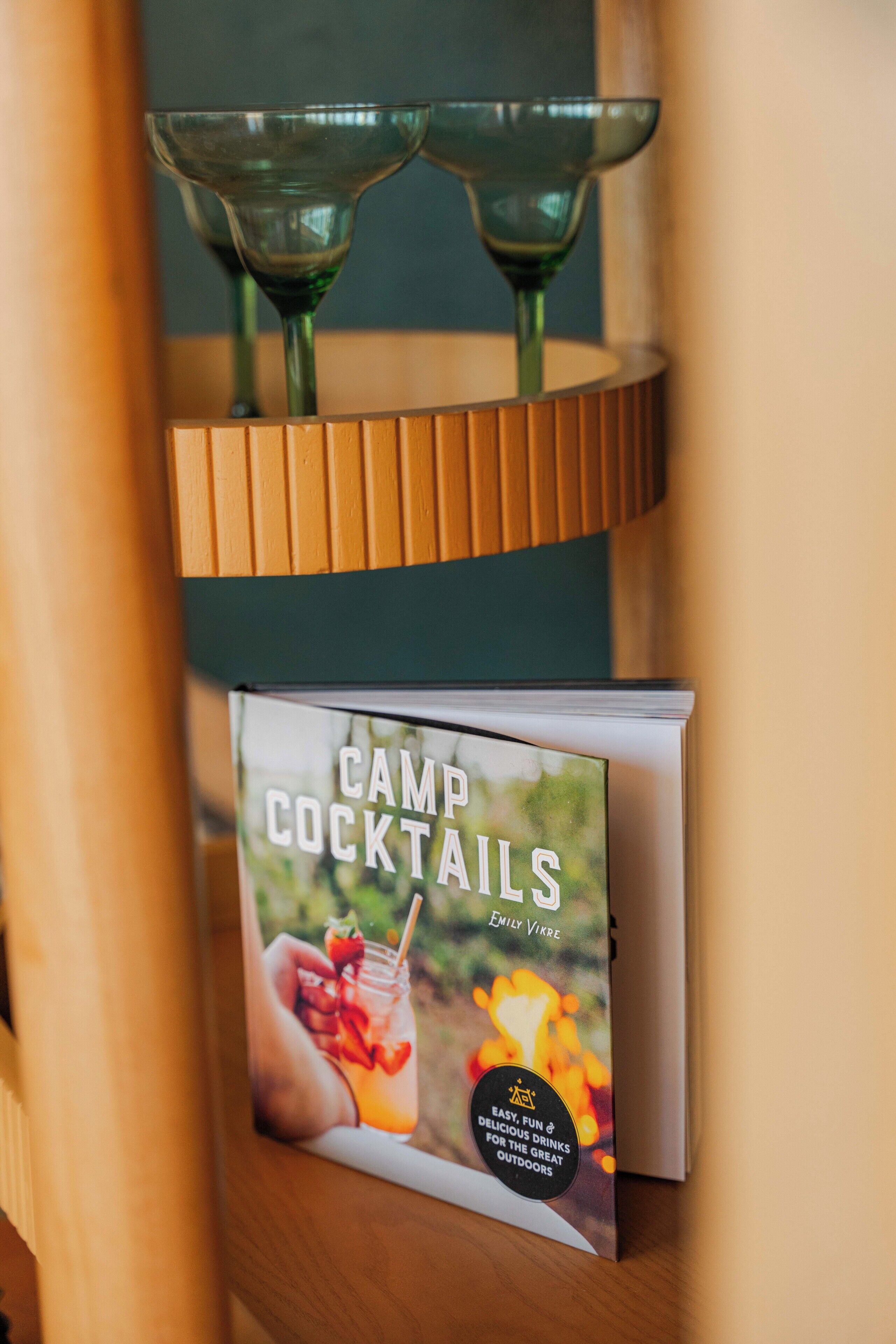 A book titled 'Camp Cocktails' by Emily Vikre on a wooden shelf.