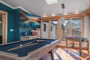 Ein Billardtisch mit Billardkugeln und Queues, ein Kicker, ein Gemälde an der Wand und ein Fenster mit Blick auf Bäume.