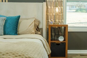 Uma cama bem arrumada com um edredom bege e almofadas, uma mesinha de cabeceira com uma garrafa de água e um pequeno alto-falante, e uma janela com cortinas estampadas.