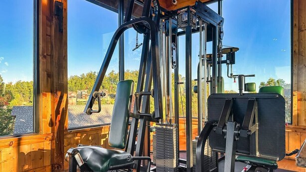 Uma academia com diversos equipamentos de ginástica, janelas amplas e vista para árvores.