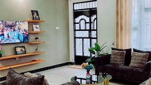 A living room with two sofas, a coffee table, and a TV on a wall. There is a door with a metal gate and a window with curtains.