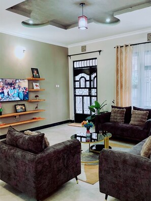 Sala de estar con dos sofás, una mesa de centro y un televisor en la pared. Hay una puerta con reja metálica y una ventana con cortinas.