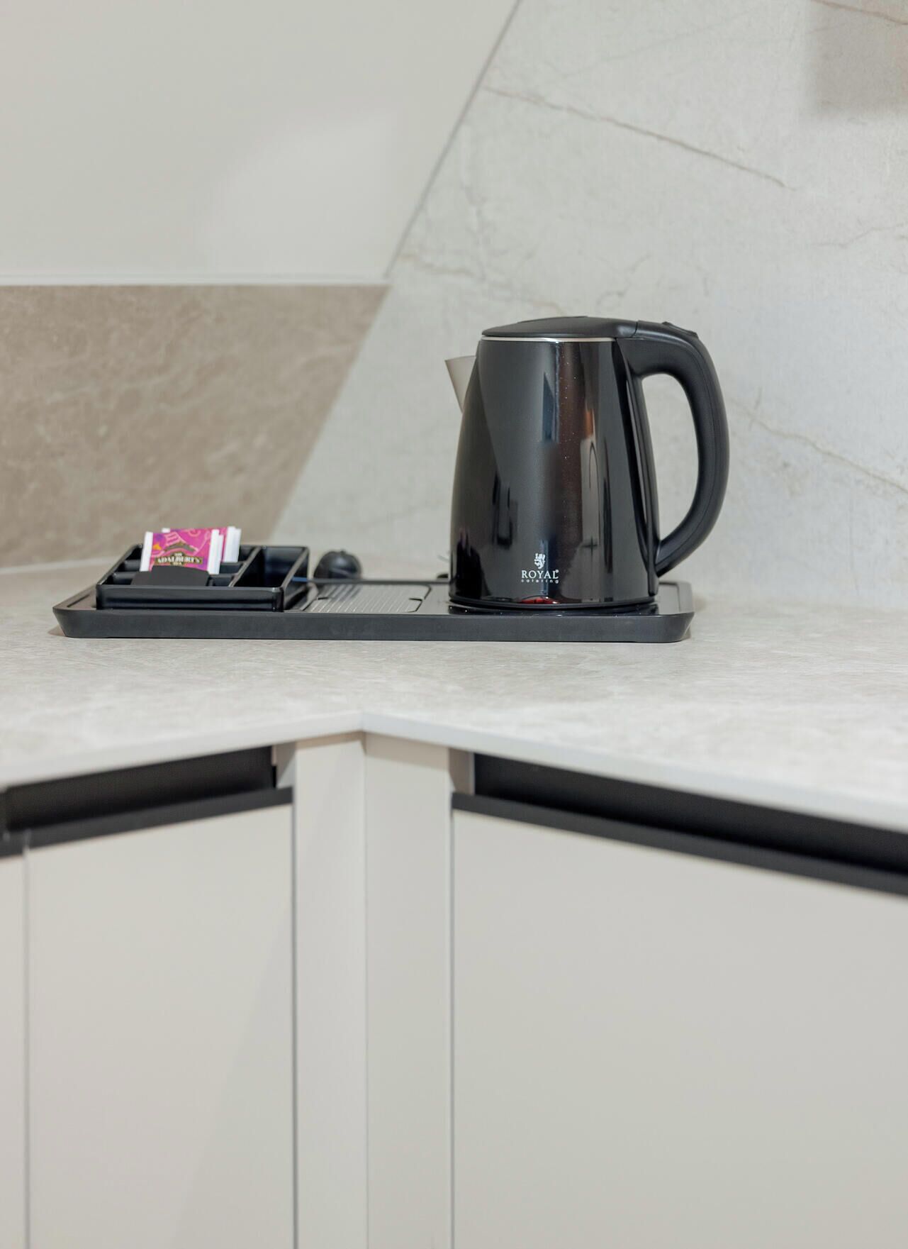 A black coffee maker on a countertop with a tray holding a pink packet and a black object, possibly a remote control.