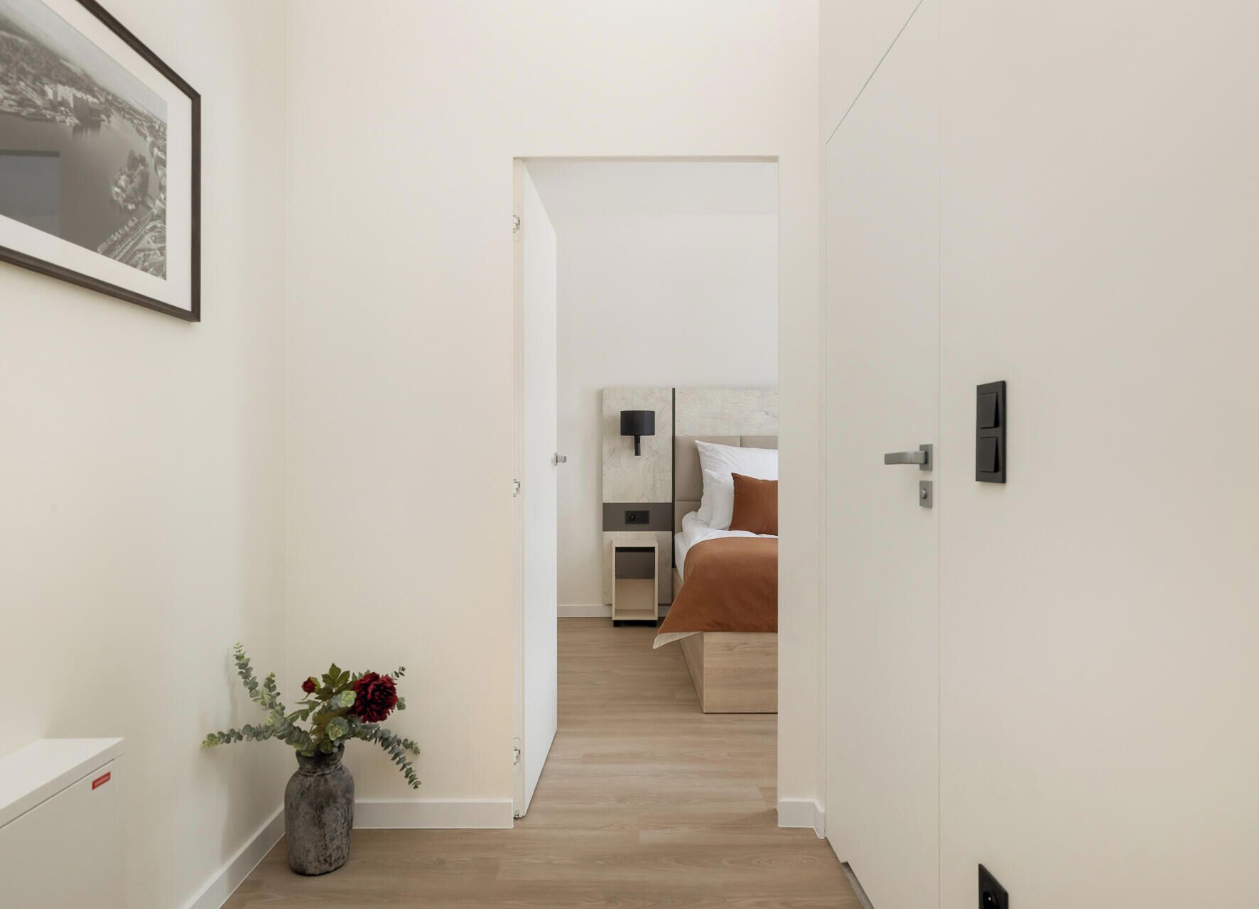 Un pasillo que conduce a un dormitorio con cama, mesita de noche y lámpara fijada a la pared.