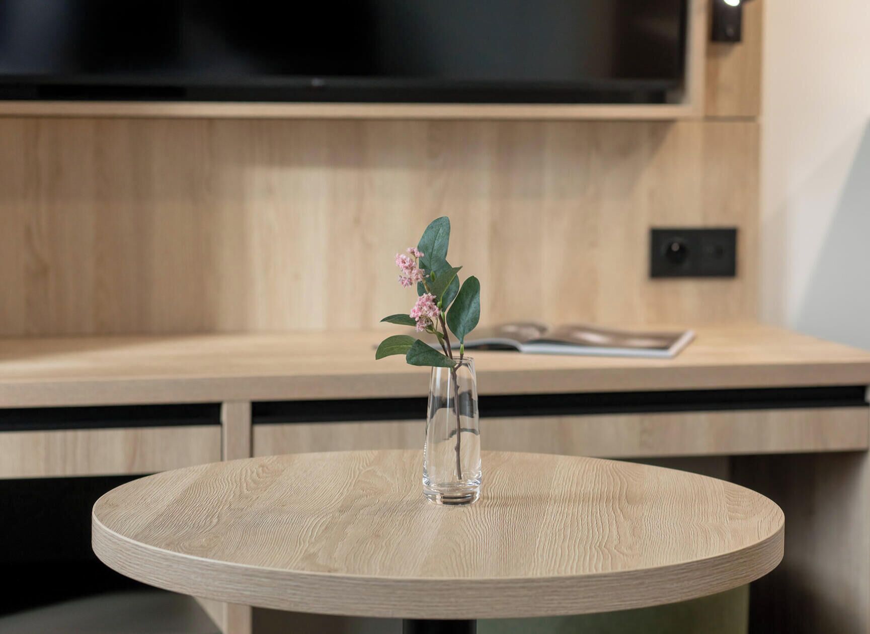 Een ronde tafel met een glazen vaas met plant en groene bladeren en roze bloemen.