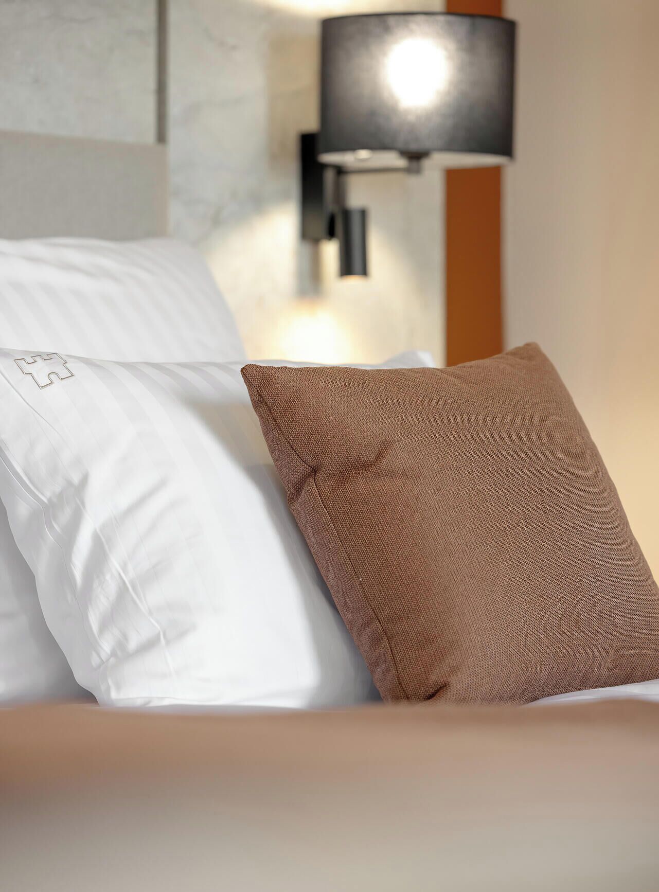 A bed with white and brown pillows, a wall-mounted lamp, and a wooden headboard.