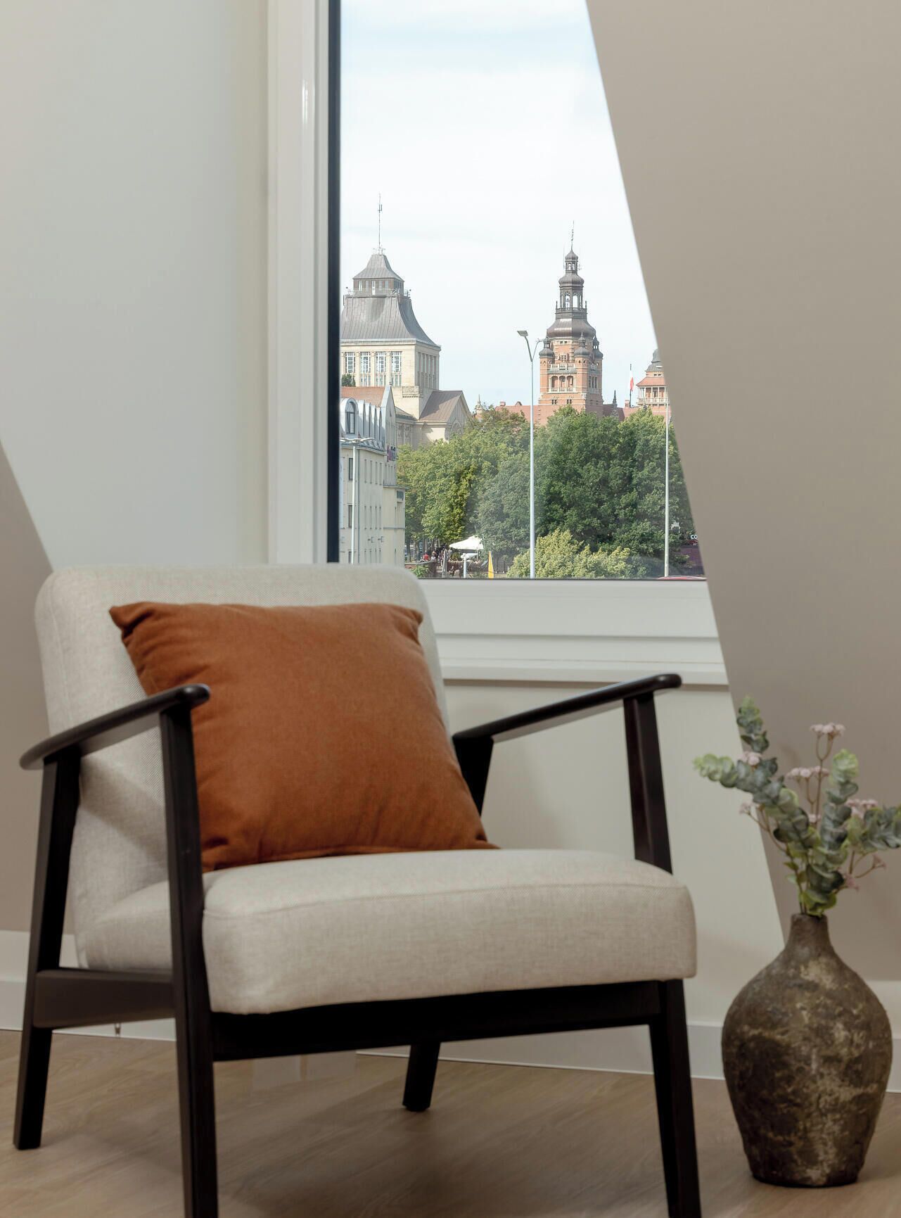 Una silla con cojín, un jarrón con flores y vista a edificios a través de una ventana.