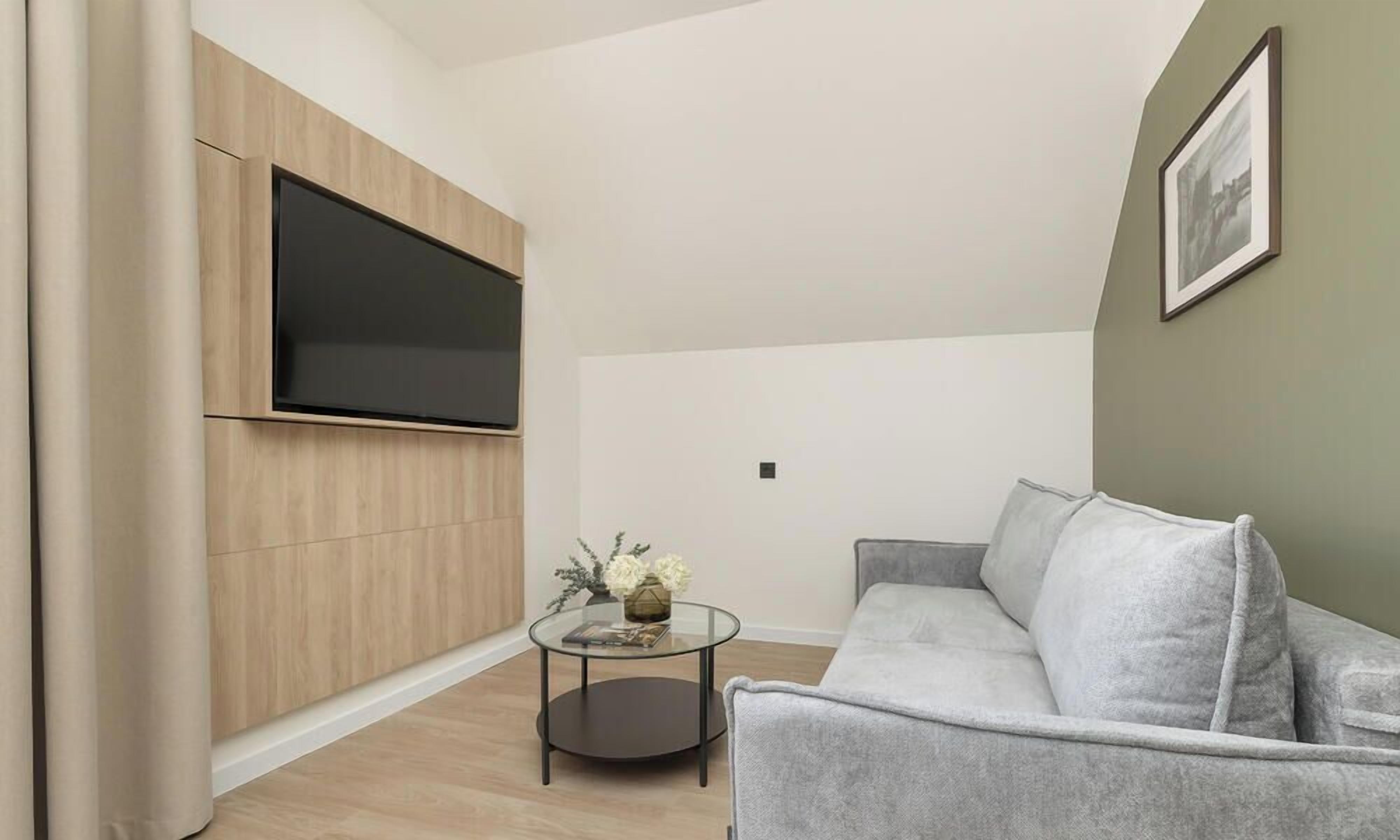 A modern living room with a flat-screen TV mounted on a wooden panel, a grey sofa, a glass coffee table with a vase and book, and a framed picture on the wall.