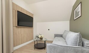 Una sala de estar moderna con un televisor de pantalla plana montado en un panel de madera, un sofá gris, una mesa de centro de vidrio con un florero y un libro, y un cuadro en la pared.