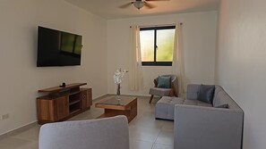 Ein modernes Wohnzimmer mit Flachbildfernseher, einer Holz-Unterhaltungseinrichtung, einem Glastisch, einem grauen Sofa und einem Fenster mit Vorhängen.