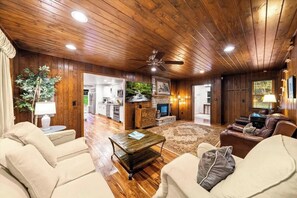 Ein geräumiges Wohnzimmer mit Holzboden, Deckenventilator, Kamin und Blick zur Küche durch eine geöffnete Tür.