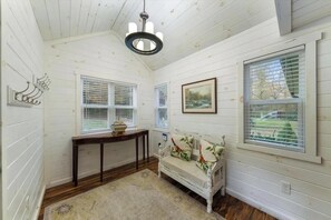 Una habitació amb parets de fusta blanca, un sofà blanc amb coixins florals, una taula consola de fusta, un penjallum i una finestra gran amb persianes.