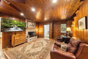 A cozy living room with a fireplace, leather sofa, and a large TV on a wooden wall. - Escape to Creek Haven, a stunning riverside retreat nestled on Norton Creek Road (Gatlinburg)
