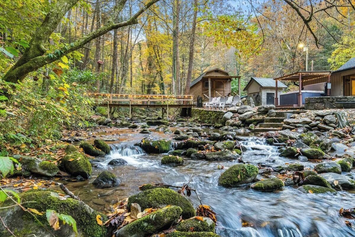 Un chalet rustique doté d’une terrasse en bois et d’une construction en pierre, entouré d’une forêt et traversé par un ruisseau.