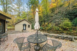 Eine Terrasse mit rundem Tisch und Stühlen, einem Sonnenschirm und einem kleinen Schuppen in einer bewaldeten Gegend.
