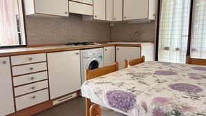 A compact kitchen with a washing machine, a dining table with floral tablecloth, wooden chairs, and a window with curtains.