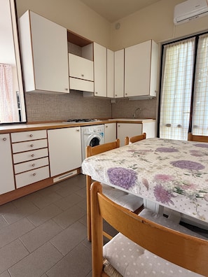 Cocina compacta con lavadora, una mesa de comedor con mantel floral, sillas de madera y una ventana con cortinas.