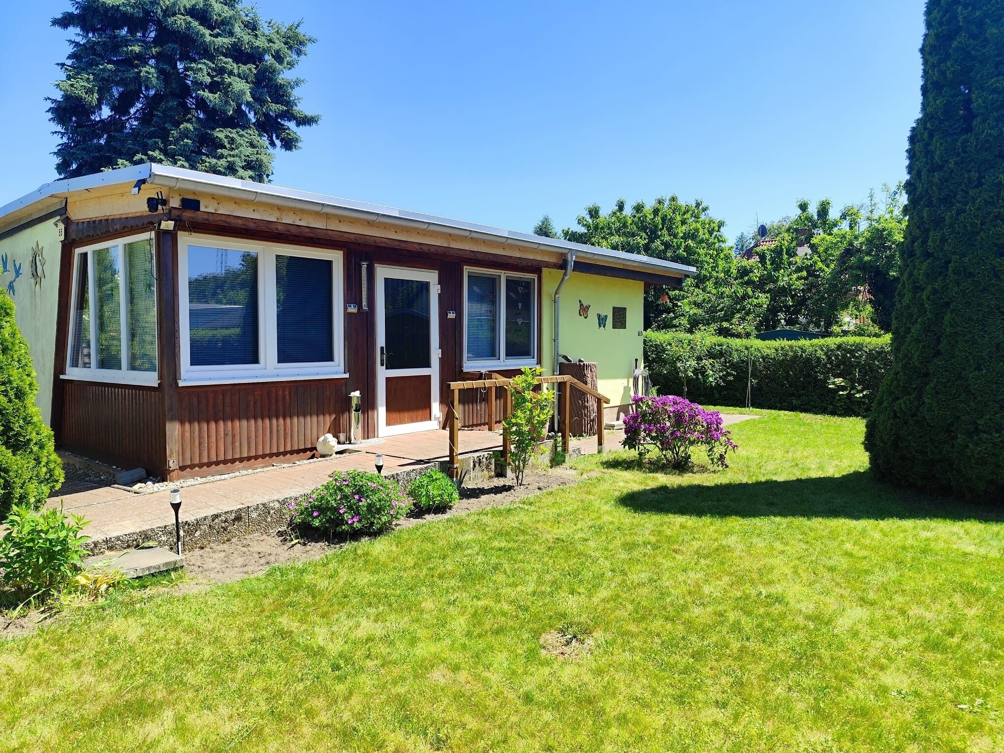 Ein einstöckiges Haus mit Veranda, ein kleiner Garten mit Blumen und einem klaren Himmel.