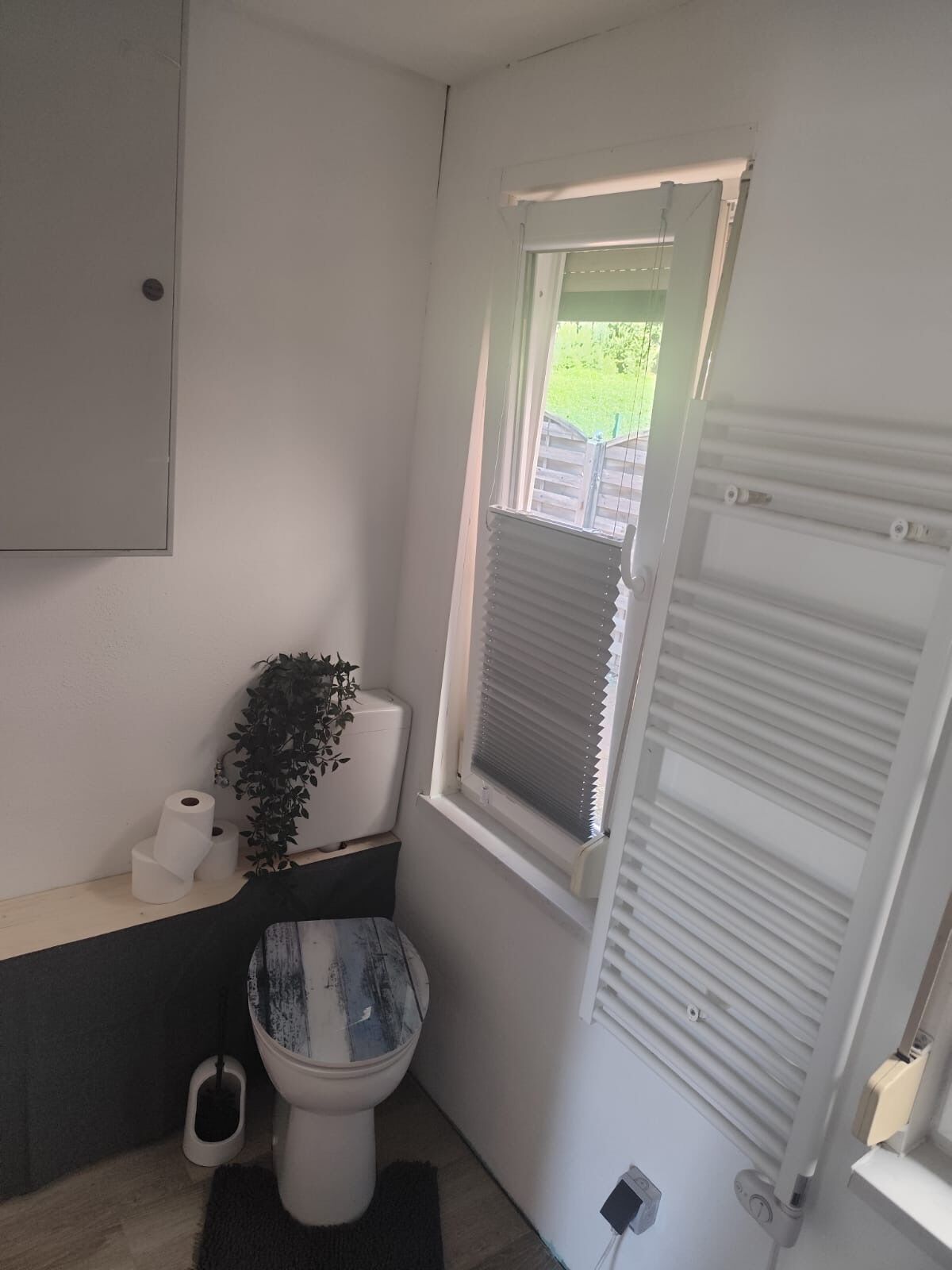 Ein kleines Badezimmer mit Toilette, einem schmallen Fenster mit Jalousien, einem Heizkörper und einer Pflanze.