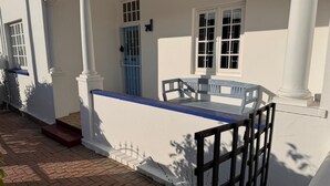 A white building with a covered porch, a blue door, and a small outdoor seating area with chairs.