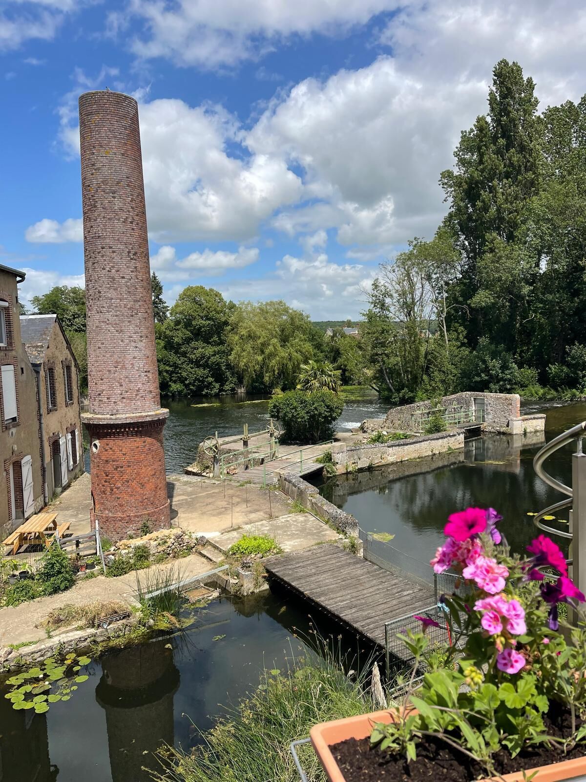 A brick chimney, a canal, and a bridge with greenery and flowers.