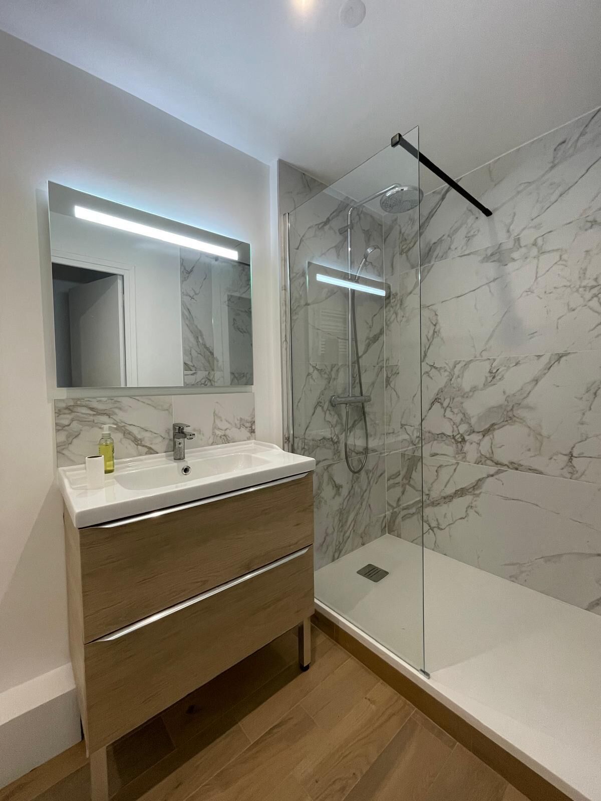 Une salle de bain avec un mur en marbre, une douche en verre et un meuble-lavabo en bois doté d’un miroir.