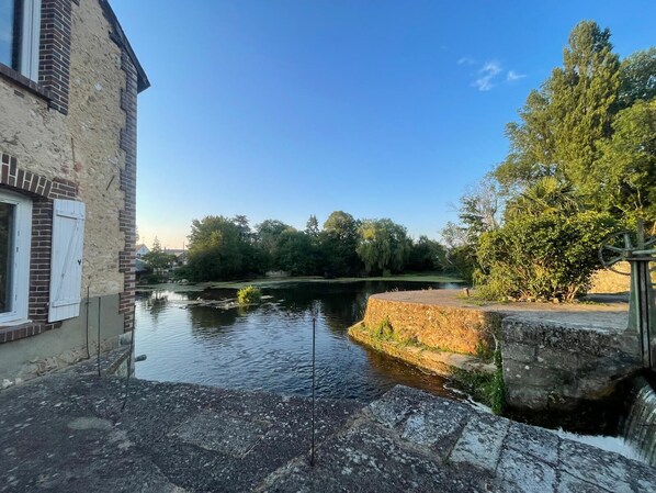 A canal with a water wheel, a stone building with a window, and trees in the background.