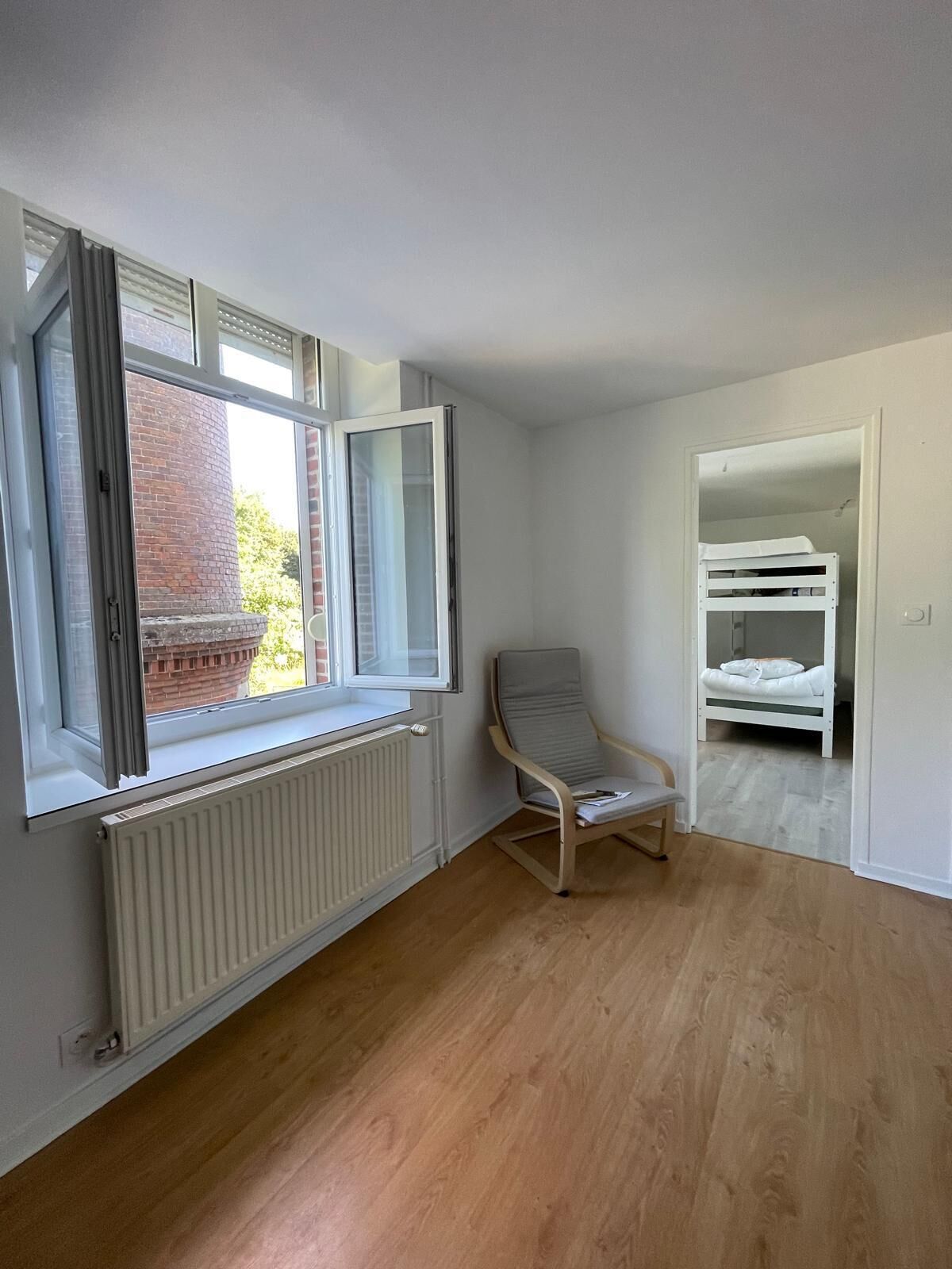 A room with a radiator, a window with open blinds, a chair, and a bunk bed in the adjacent room.