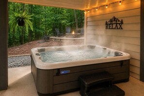 A hot tub with a control panel, surrounded by a wooden deck and a stone fire pit, with a view of a forested area. - River Resort Retreat | Hot Tub & Game Room | Pets (Ellijay)