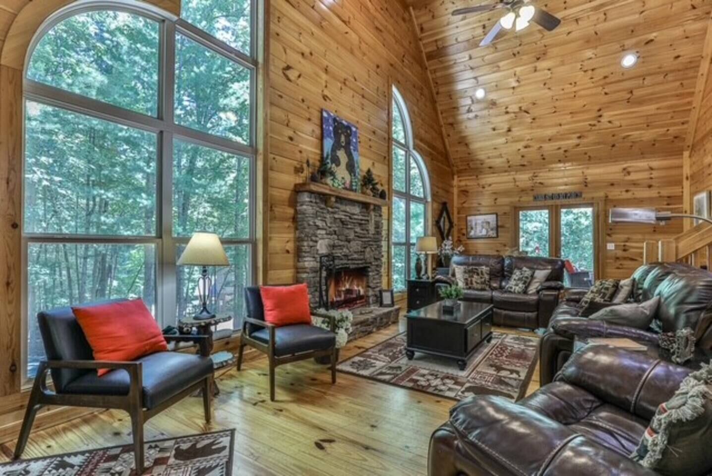 A log cabin interior with a stone fireplace, leather sofas, and large windows offering a forest view.