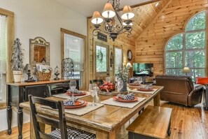 A dining area with a wooden table set for four, surrounded by chairs, a chandelier, and a television in the background. - River Resort Retreat | Hot Tub & Game Room | Pets (Ellijay)