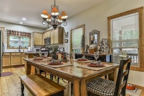 Un coin repas avec une table en bois dressée pour quatre personnes, entourée de chaises dotées de coussins à carreaux. Un lustre est suspendu au-dessus de la table, et la pièce est équipée de meubles en bois et d’une fenêtre avec des stores.