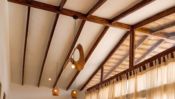 Une pièce avec un plafond haut, des poutres en bois, une chaise en osier, un coin cuisine et une table à manger.