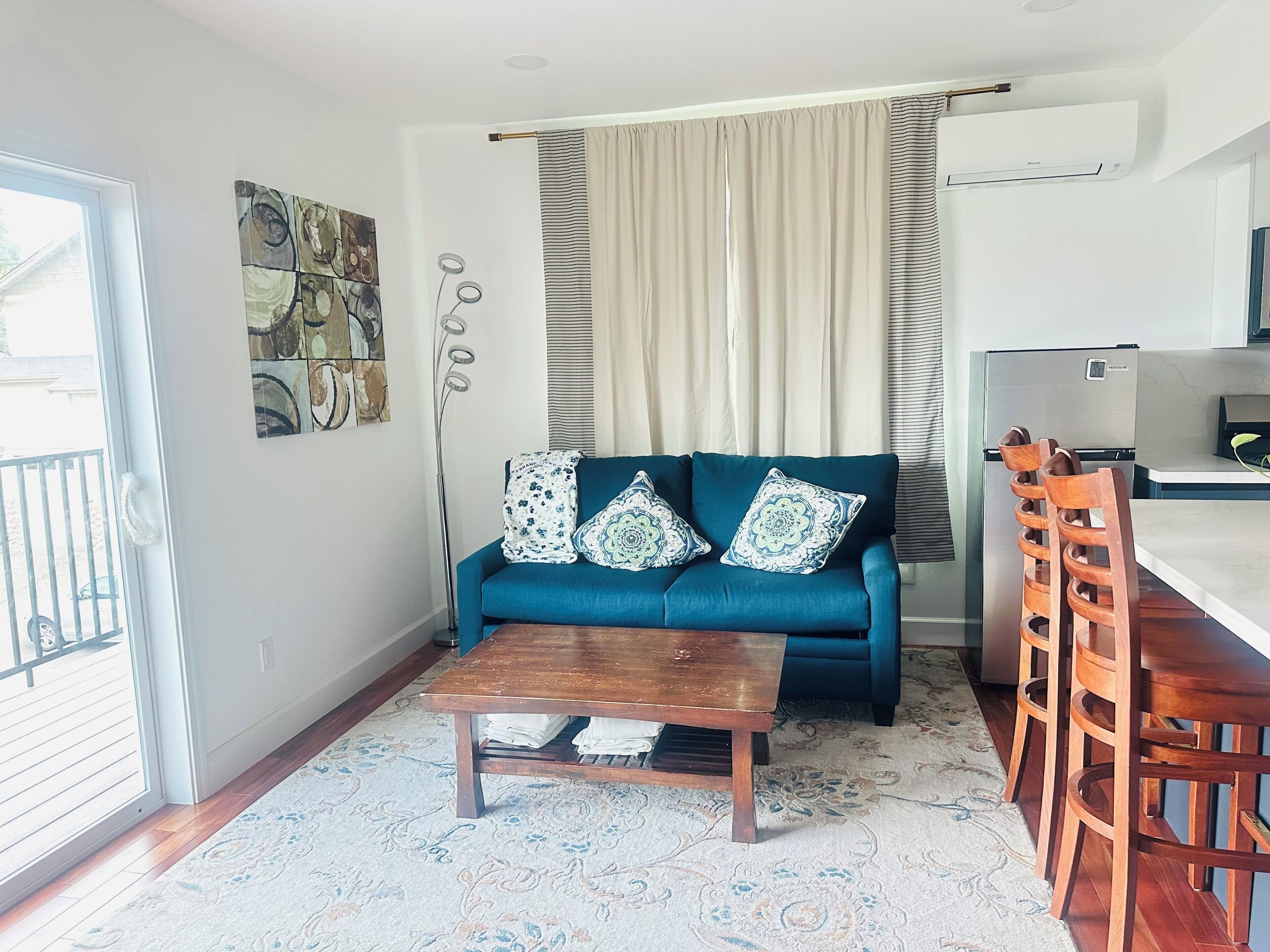 A living room with a sofa, a coffee table, a dining table with chairs, a kitchen area, and a large window with curtains.