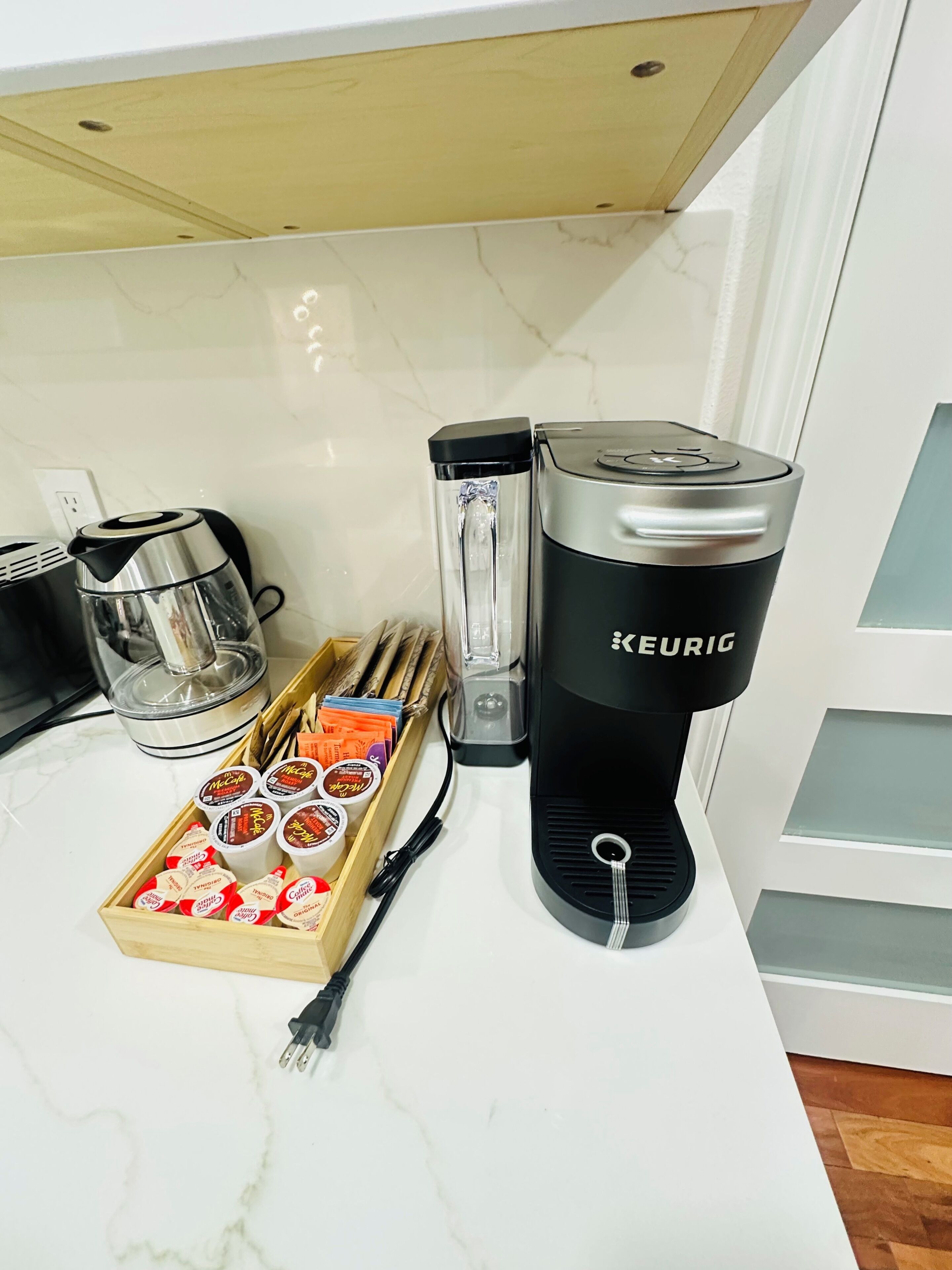 Un comptoir de cuisine avec une machine à café Keurig, une verseuse en verre, un plateau en bois contenant des dosettes McCafé et une bouilloire électrique noire.