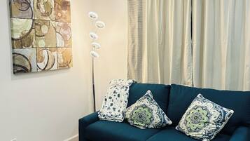 A living room with a blue sofa, two patterned cushions, a coffee table, a wall art piece, and a window with curtains.