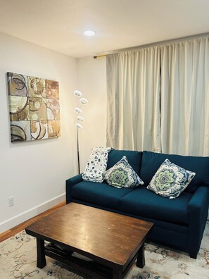 A living room with a blue sofa, two patterned cushions, a coffee table, a wall art piece, and a window with curtains. - Luxury Multi-Level Suite with Deck & Mountain View (Renton)