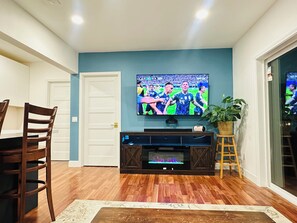 A living room with a flat-screen TV mounted on the wall, a wooden entertainment unit, a dining table with chairs, and a potted plant. - Luxury Multi-Level Suite with Deck & Mountain View (Renton)